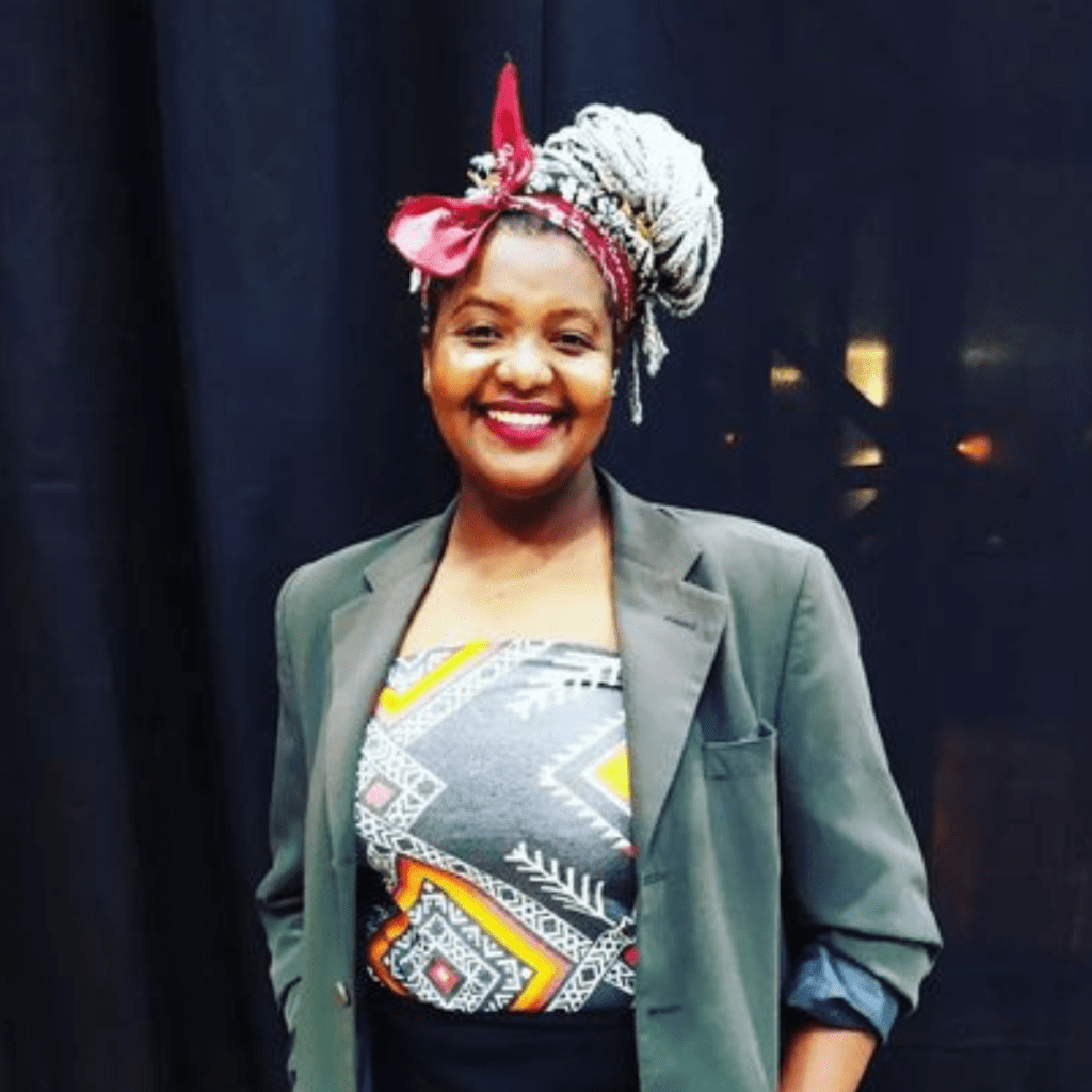 fotografia de Fátima Tertuliano. Mulher de pele negra com um lenço amarrado sobre os cabelos, veste um blazer cinza sobre uma camisete estampada.