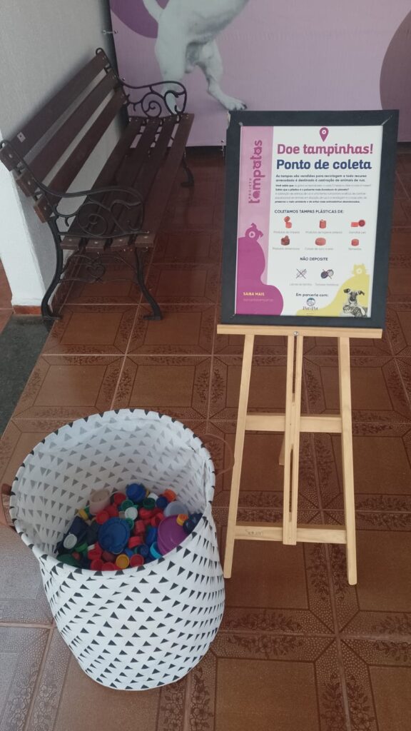 Bacia com tampas plásticas ao lado de um banner escrito "Doe tampinhas! Ponto de coleta" e com informações sobre o Projeto Tampatas. Ao fundo tem um banco de madeira e uma parede rosa com a imagem de um cachorro.